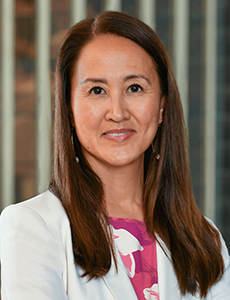 A woman with long brown hair wearing a white blazer over a pink patterned blouse stands in front of a window. She is smiling and looking at the camera.