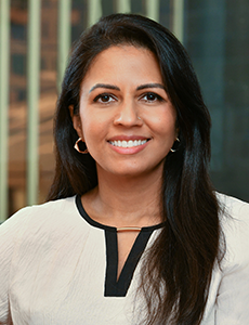 A woman with long dark hair and hoop earrings, wearing a cream-colored blouse with black trim, smiles at the camera. The background shows vertical blinds and blurred office windows.